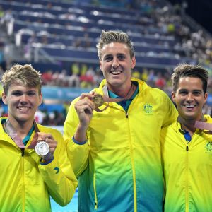 Image of 3 Australian Para-swimmers dressed in gold tracksuits, smiling at the camera and holding medals in their left handss.