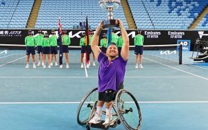 Australian wheelchair tennis player Dylan Alcott smiling at the camera and lifting a trophy above his head
