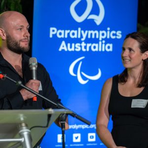 Image of male standing holding a microphone in his right hand speaking and a female standing to his left. They are both standing in front of a blue backdrop with the Paralympics Australia logo