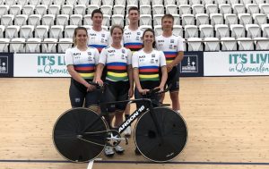 Image of a group of 6 cyclists in their racing suits, standing on the velodrome track with a bike in front of them. They are smiling at the camera.
