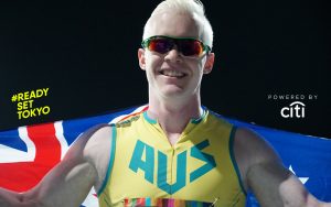 Image of Australian Paralympian Chad Perris wearing a green and gold singlet with AUS acrossthe chest in green letters. He is smiling at the camera and wearing tinted glasses, and holding the Australian flag around his shoulders.