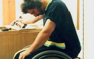 Image of a male in a wheelchair with his head looking down. He is wearing ear muffs and glasses and holds a pistol in his right hand preparing to shoot in a competition. His wheelchair has the words Australia in green and gold on the wheel.