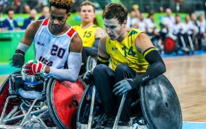 Image of two males playing wheelchair rugby contesting the ball. The male on the left is wearing a USA uniform, the male on the right is wearing an Australian uniform
