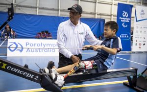 A young male left leg amputee on a rowing machine being supervised by an adult
