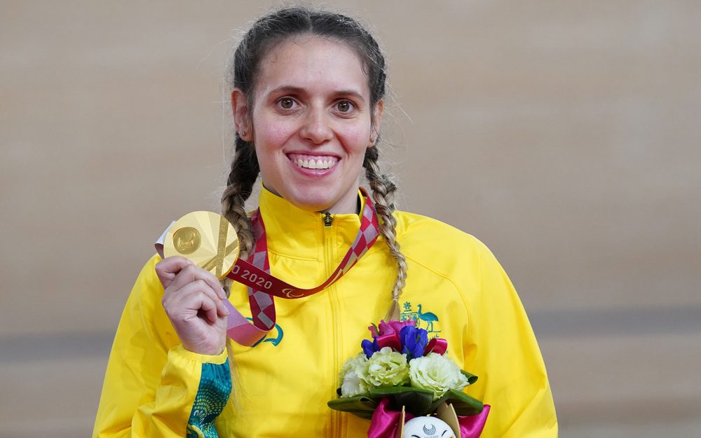 Smiling female wearing a yellow jacket holding a gold medal in her right hand.