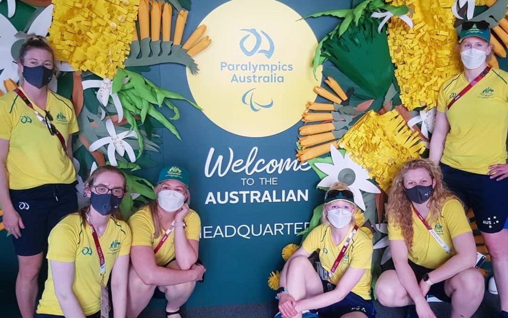 Members of the Australian Belles Goalball team pose for a photo in front of a green wall covered in Australian native plants.