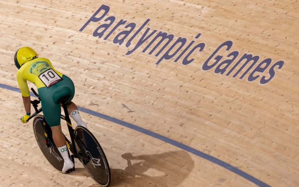 A track cyclist wearing a green and gold racing suit is cycling at a velodrome