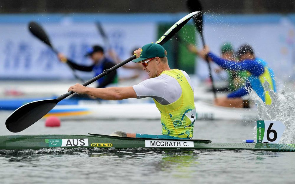 Australian Paralympian Curtis McGrath is wearing a yellow singlet over a white shirt, ad a green hat. He is paddling in his green kayak competing at the Tokyo 2020 Paralympic Games