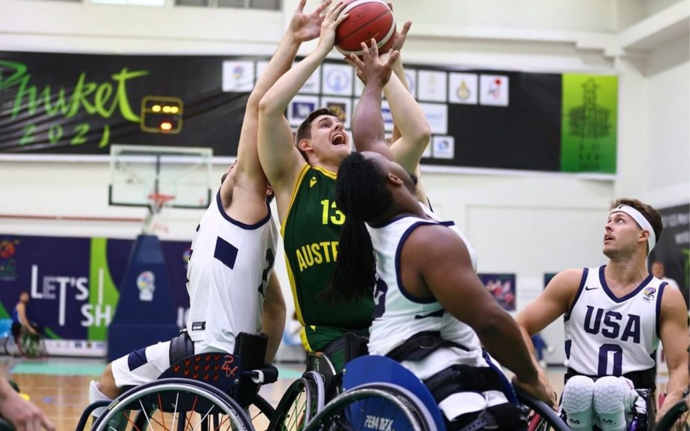 An Australian U23 Wheelchair basketball player in a green singlet taking a shot. He is surrounded by defenders from the other team.