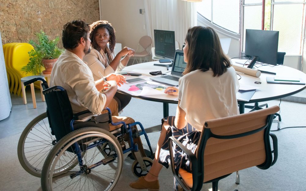 A group of three professionals sit around a circular table discussing information on a computer screen.