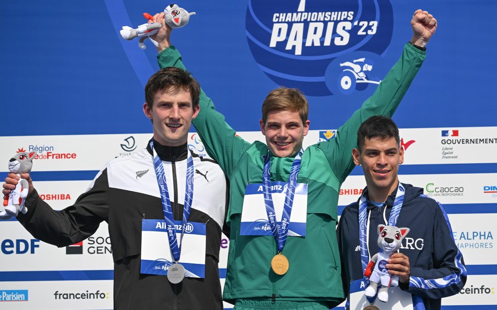 Three athletes on a podium. In the middle is Australian Paralympian James Turner smiling and raising both hands in the air in celebration.