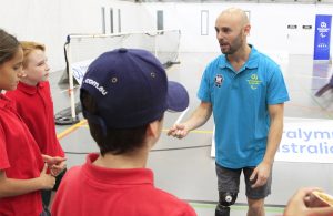 Image of Australian male Paralympian in a blue t-shirt talking to a group of 3 school children.